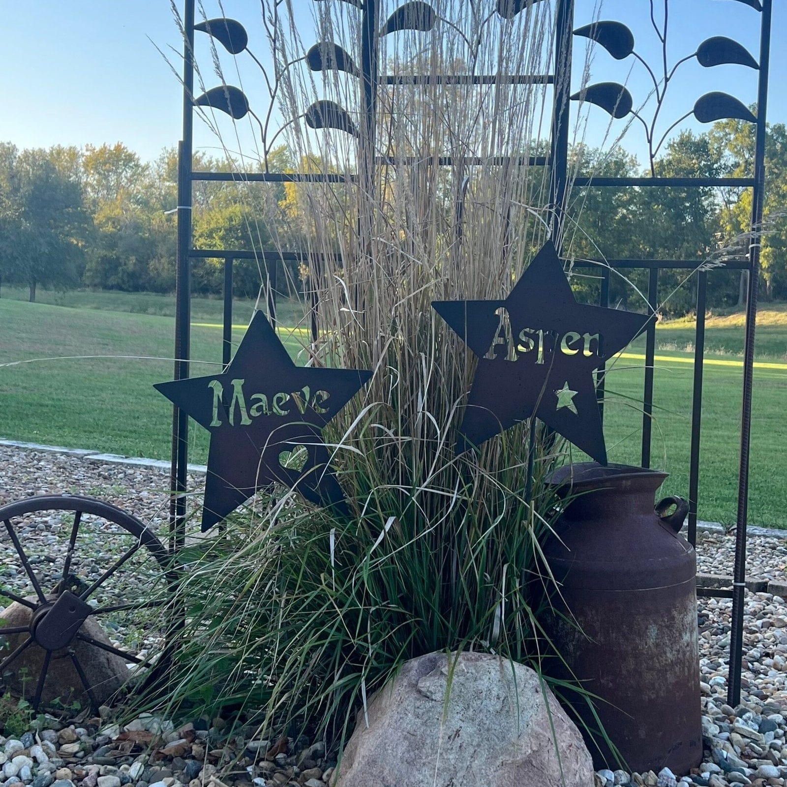 Two Handcrafted Metal Name Stars – Personalized Outdoor Garden Art, custom-labeled Maeve and Aspen, are displayed in tall grass among a milk can, wagon wheel, and stones with a green lawn and trees in the background.