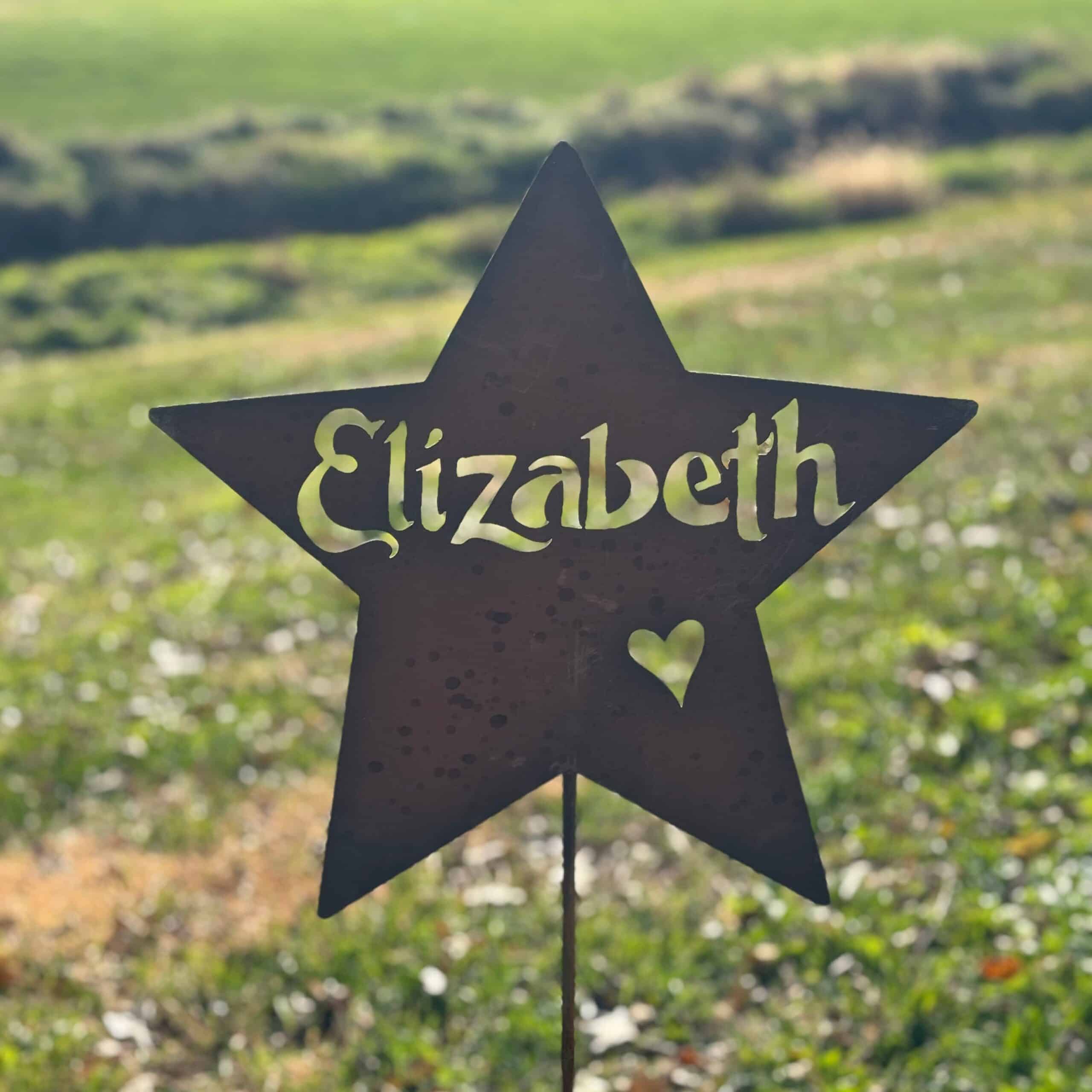 The Handcrafted Metal Name Star – Personalized Outdoor Garden Art, featuring "Elizabeth" and a heart cutout, stands in a grassy field with sunlight softly illuminating the background.