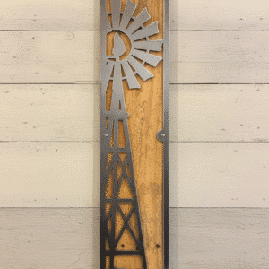 A metal silhouette of a windmill mounted on a rectangular wooden plaque, hung vertically on a light-colored wooden plank wall.