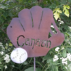 The Baseball Glove Garden Stake features a metal glove shape with "Canaan" cut out and a small baseball, adding unique baseball decor to your garden among green leaves and white wildflowers.