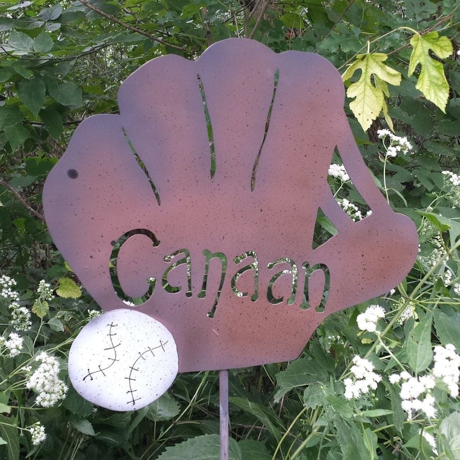 The Baseball Glove Garden Stake features a metal glove shape with "Canaan" cut out and a small baseball, adding unique baseball decor to your garden among green leaves and white wildflowers.