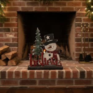 A fireplace with a brick surround is decorated with the Custom Holiday Sign Board – Mix & Match Snowman Set, featuring a snowman in a black hat, a Christmas tree, and "believe" in red. Firewood and tools sit on either side.