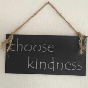 The Choose Kindness Metal Sign is a black rectangular metal wall decor featuring "choose kindness" in white lowercase letters, hung by brown twine on a white textured wall.