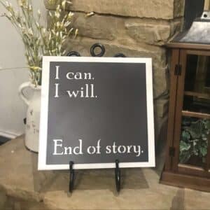 The I Can I Will Metal Sign, featuring bold white text on black metal, rests on a stone surface beside white flowers and a wooden cabinet with green foliage.