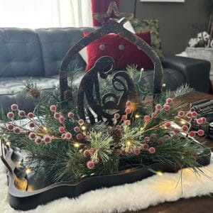 The Handcrafted Metal Nativity Tabletop Décor features Mary, Joseph, and baby Jesus on a wooden tray, accented with pine branches, red berries, and lights—an ideal centerpiece for adding cozy charm to your living room.