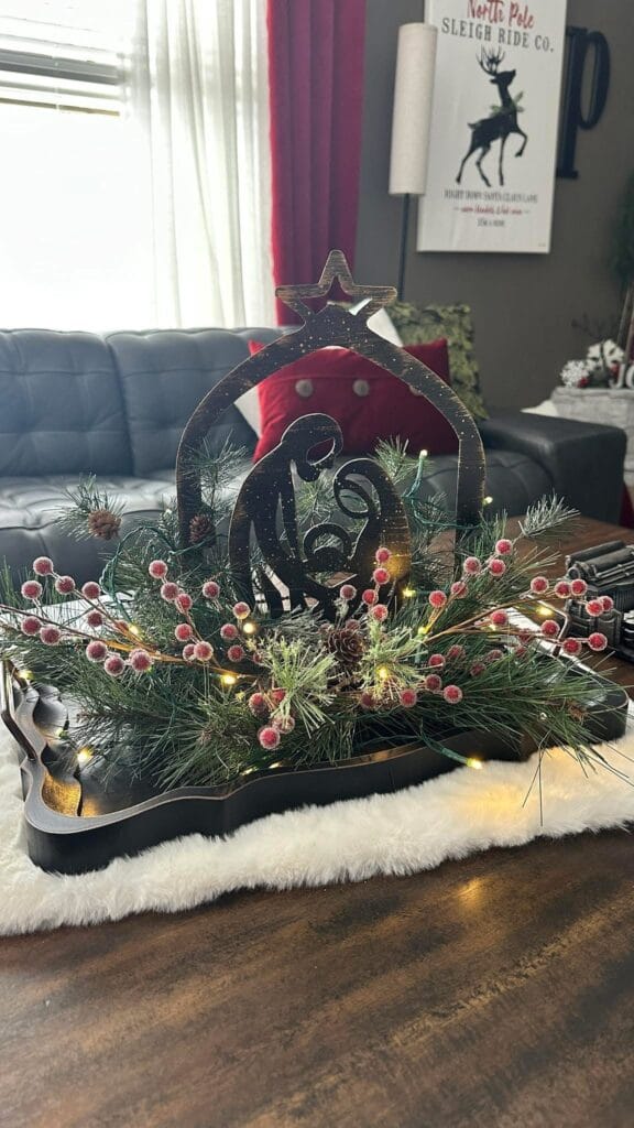 The Handcrafted Metal Nativity Tabletop Décor features Mary, Joseph, and baby Jesus on a wooden tray, accented with pine branches, red berries, and lights—an ideal centerpiece for adding cozy charm to your living room.