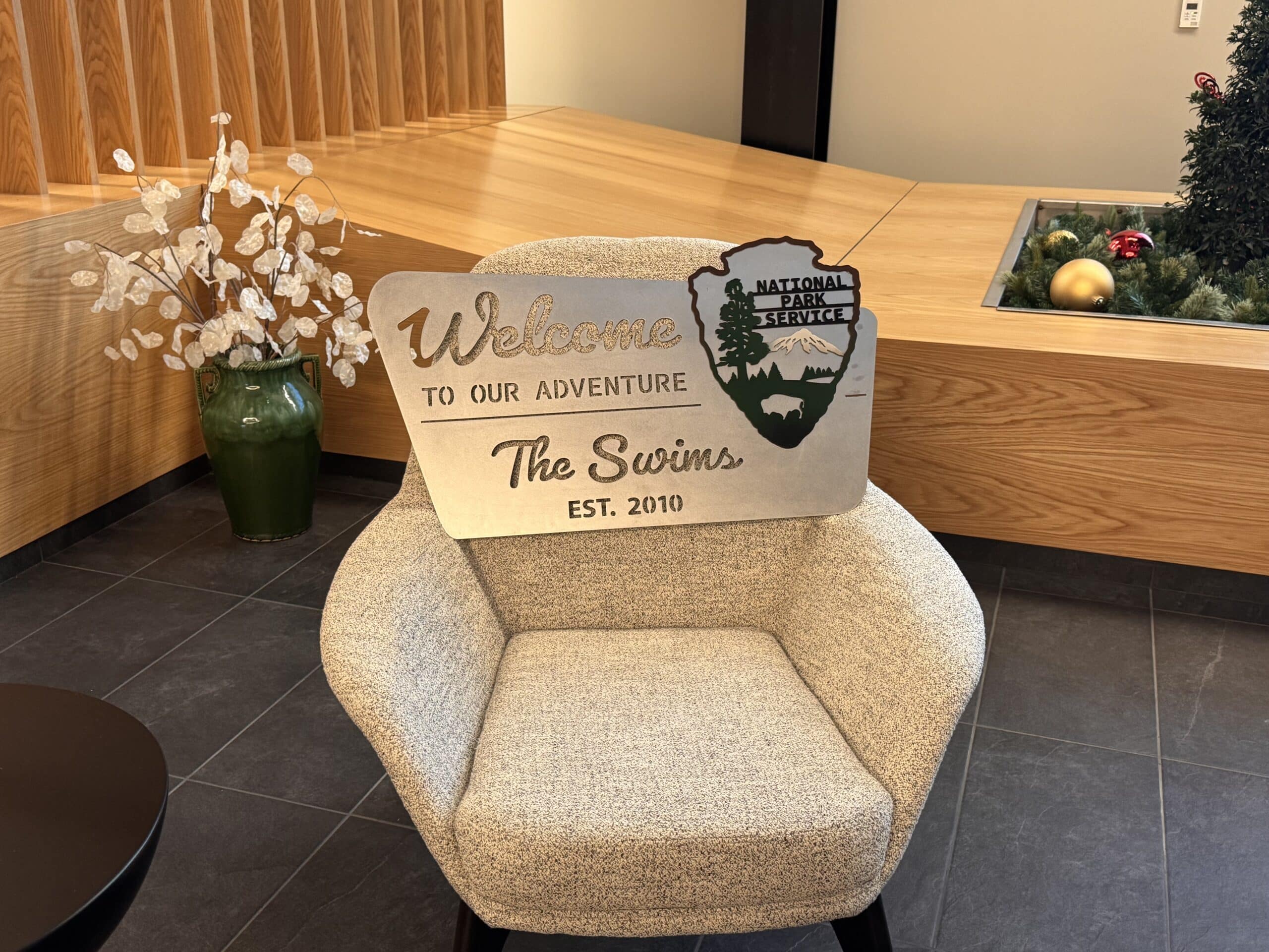 A beige cushioned chair holds the Unique Personalized National Park Sign - Custom Decor featuring “Welcome to our adventure, The Swins, Est. 2010” with tree and mountain silhouettes; white flowers and holiday decor appear in the background.