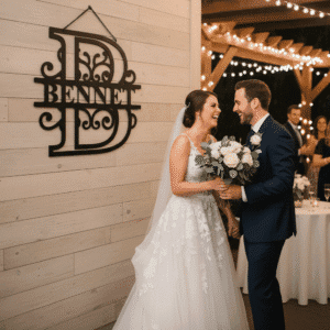 A bride and groom smile and hold hands at their wedding reception beside a monogrammed Bennet sign, with string lights and guests in the background. The bride holds a bouquet of white flowers.